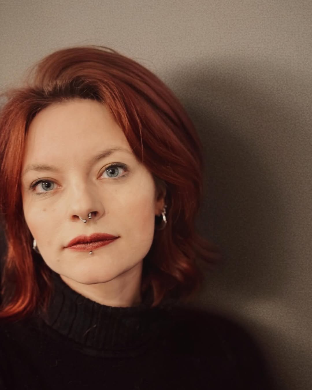 Elisa Garrett Portrait von Elisa Garrett mit kupferrotem Haar, hellem Teint und Piercings an Nase und Unterlippe. Sie trägt einen schwarzen Rollkragenpullover und blickt ruhig in die Kamera vor neutralem Hintergrund.